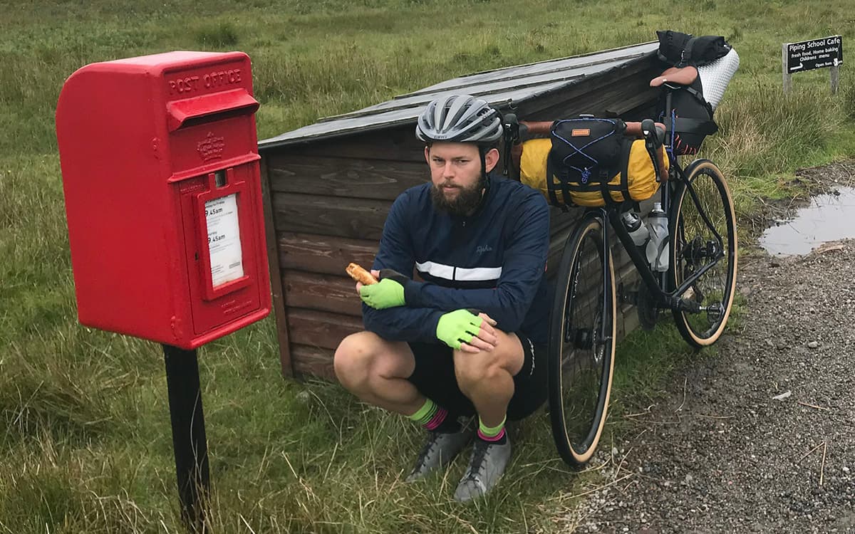 Man with a bicycle and backpack sitting next to a red postbox in a rural setting